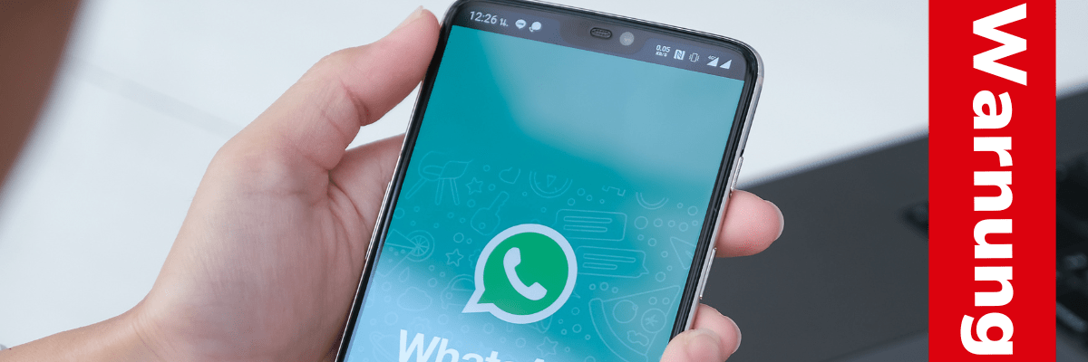 Jemand hält ein Smartphone, auf dem Display ist das WhatsApp-Logo zu sehen. Auf einem roten Ausrufezeichen steht "Warnung".